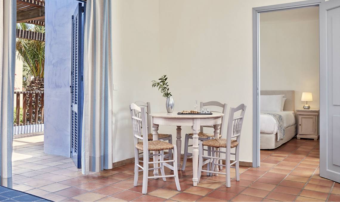 Inneneinrichtung eines Zimmers im Candia Park Village Hotel mit Tisch, Stühlen und einem Blick auf einen Balkon und ein Bett.