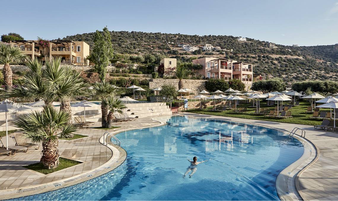 Person schwimmt im Pool des Candia Park Village Hotels, umgeben von Gebäuden und Landschaft.
