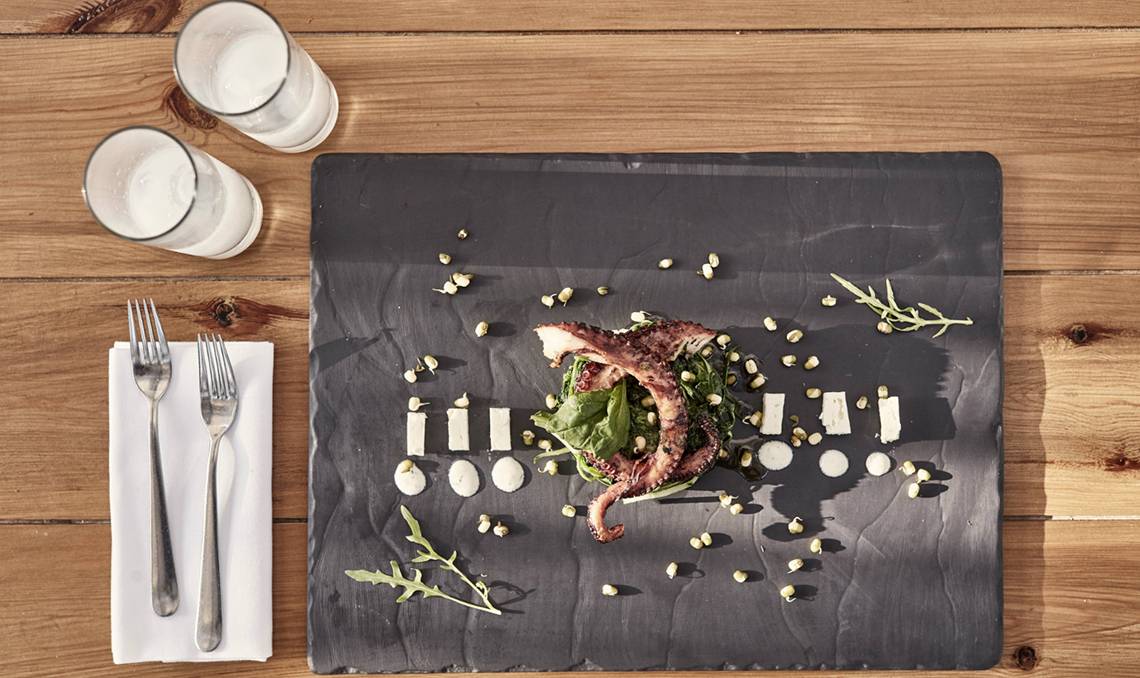 Gegrillter Tintenfisch mit Salat auf einem schwarzen Teller auf einem Holztisch mit zwei Gläsern Wasser, Gabeln und Servietten.