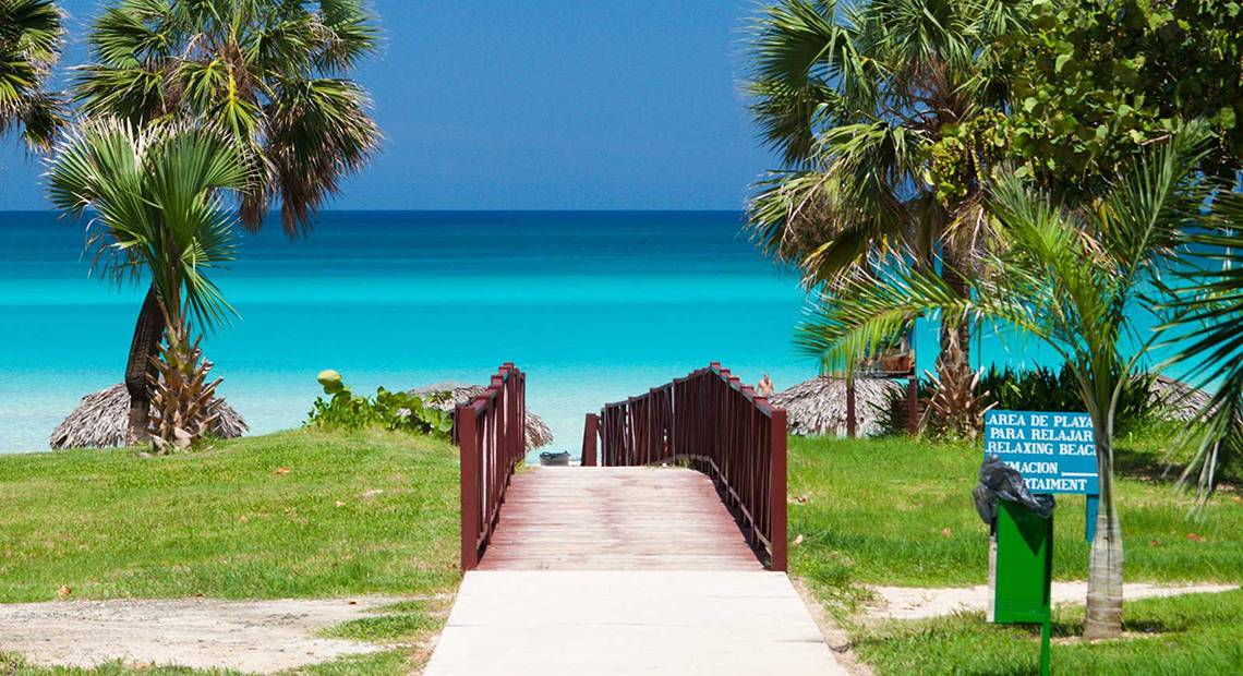 Eine Holzbrücke führt vom Hotel Occidental Arenas Blancas zum Strand mit türkisfarbenem Wasser.