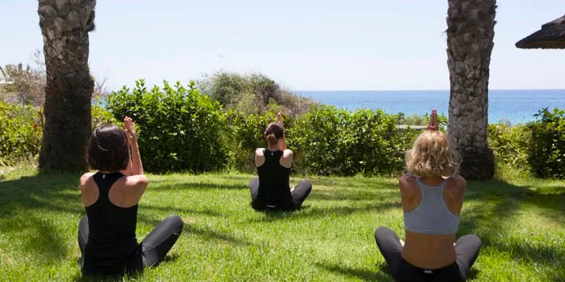 Drei Frauen praktizieren Yoga im Garten des Alion Beach Hotels mit Blick auf das Meer.