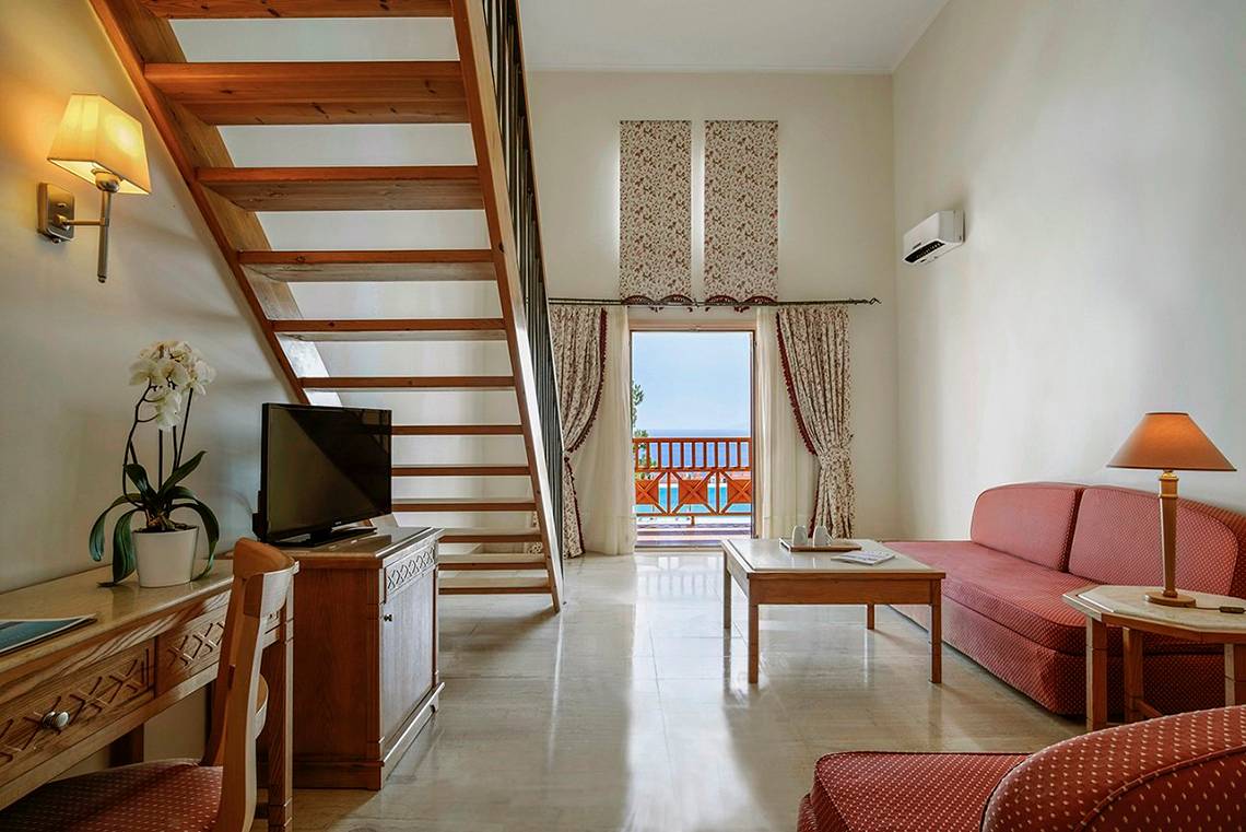 Innenansicht des Wohnzimmers im Mitsis Family Village Beach Hotel mit Sofa, Tisch, Fernseher und Balkon