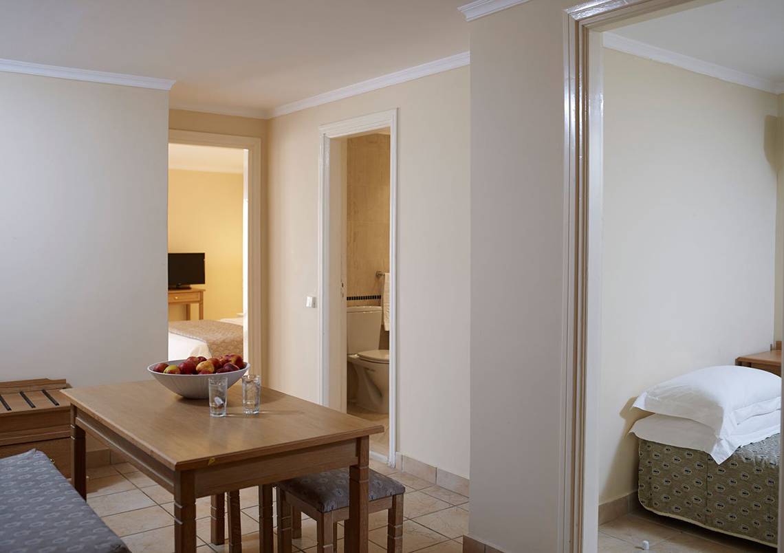 Innenansicht eines Zimmers im Mitsis Family Village Beach Hotel mit Tisch, Obstschale und Schlafzimmer.
