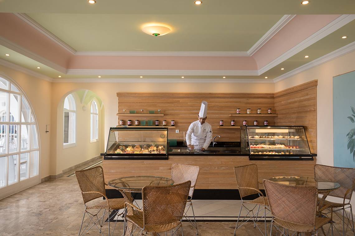 Innenansicht des Restaurants des Mitsis Family Village Beach Hotels mit einem Koch am Tresen, umgeben von Tisch und Stühlen.
