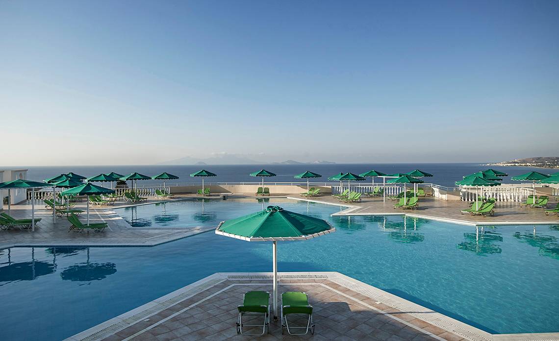 Außenpoolbereich des Mitsis Family Village Beach Hotels mit grünen Sonnenschirmen und Liegestühlen am Meer
