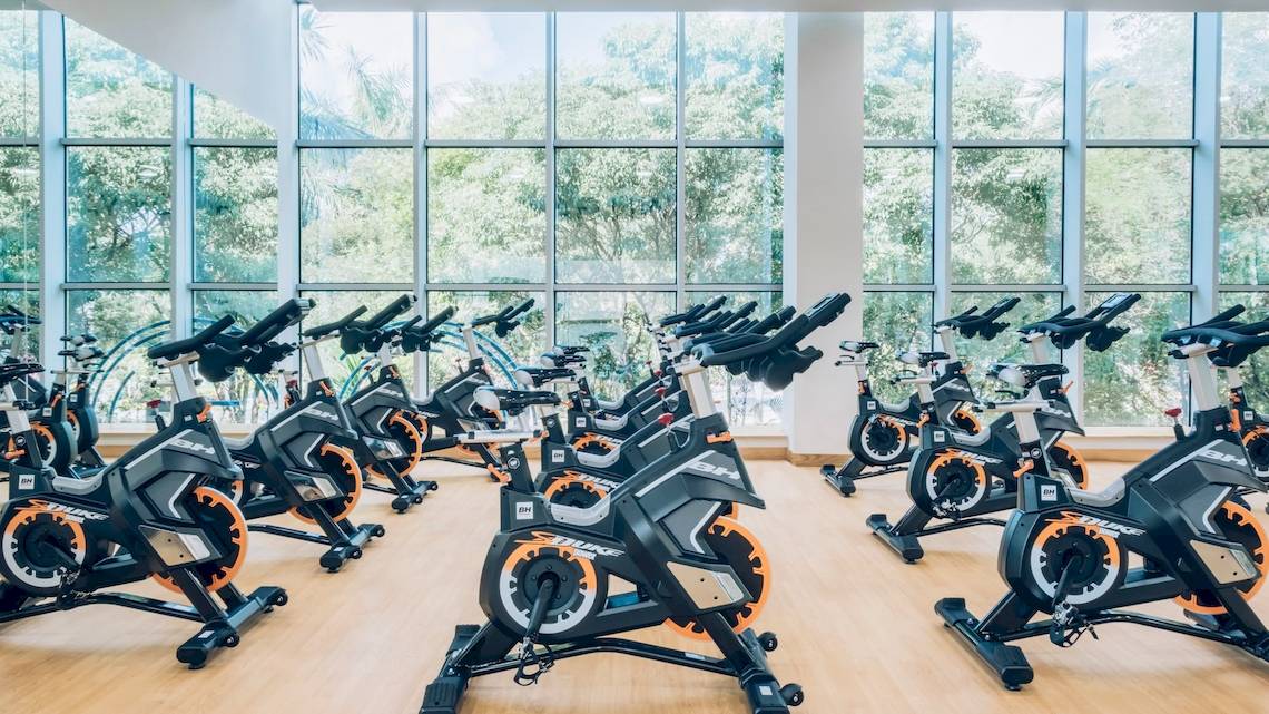 Fitnessstudio mit mehreren Fahrradtrainern vor großen Fenstern mit Blick auf Bäume