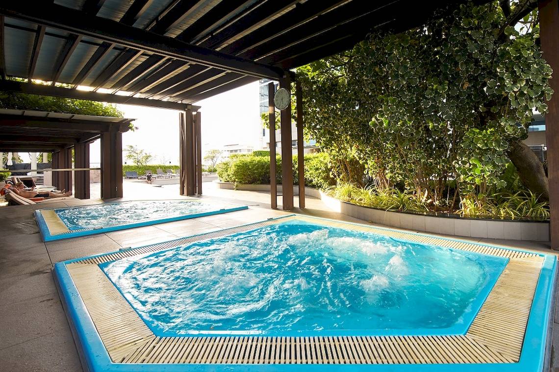 Zwei blaue Pools unter einem Dach im Pathumwan Princess Hotel mit Pflanzen und Sonnenliegen.