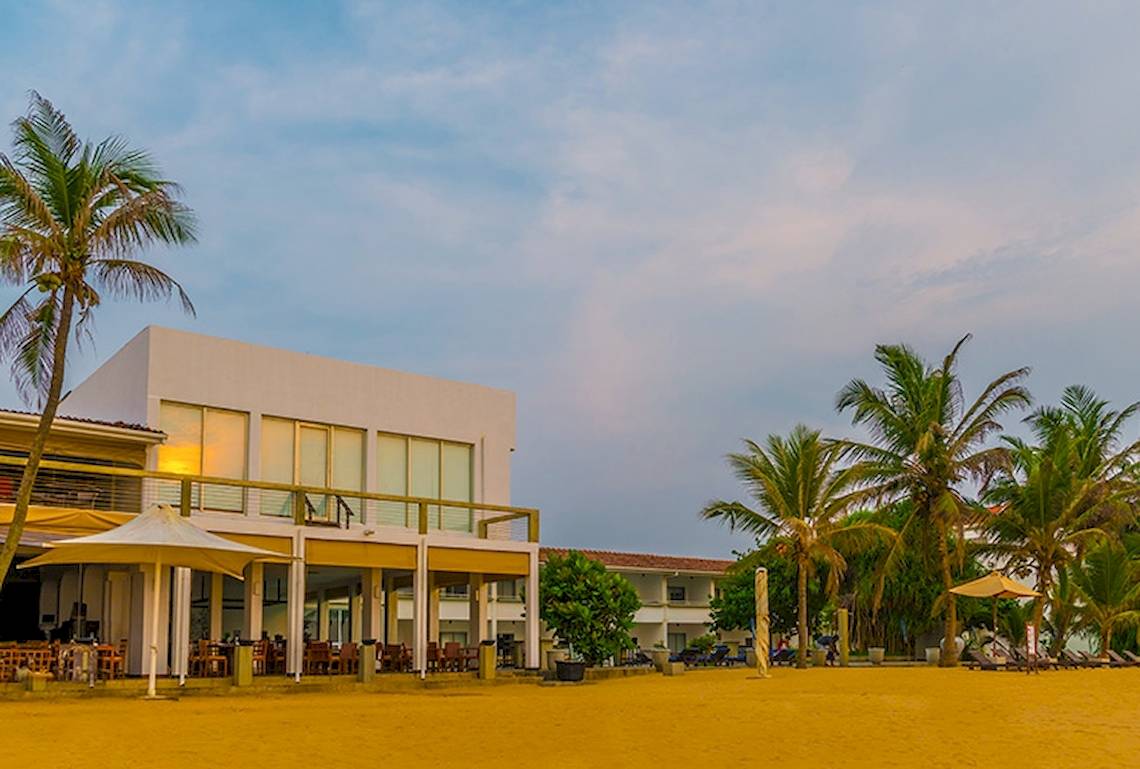 Außenansicht des Jetwing Sea Hotels mit weißem Gebäude, Terrassen, Sonnenschirmen und Palmen am Strand
