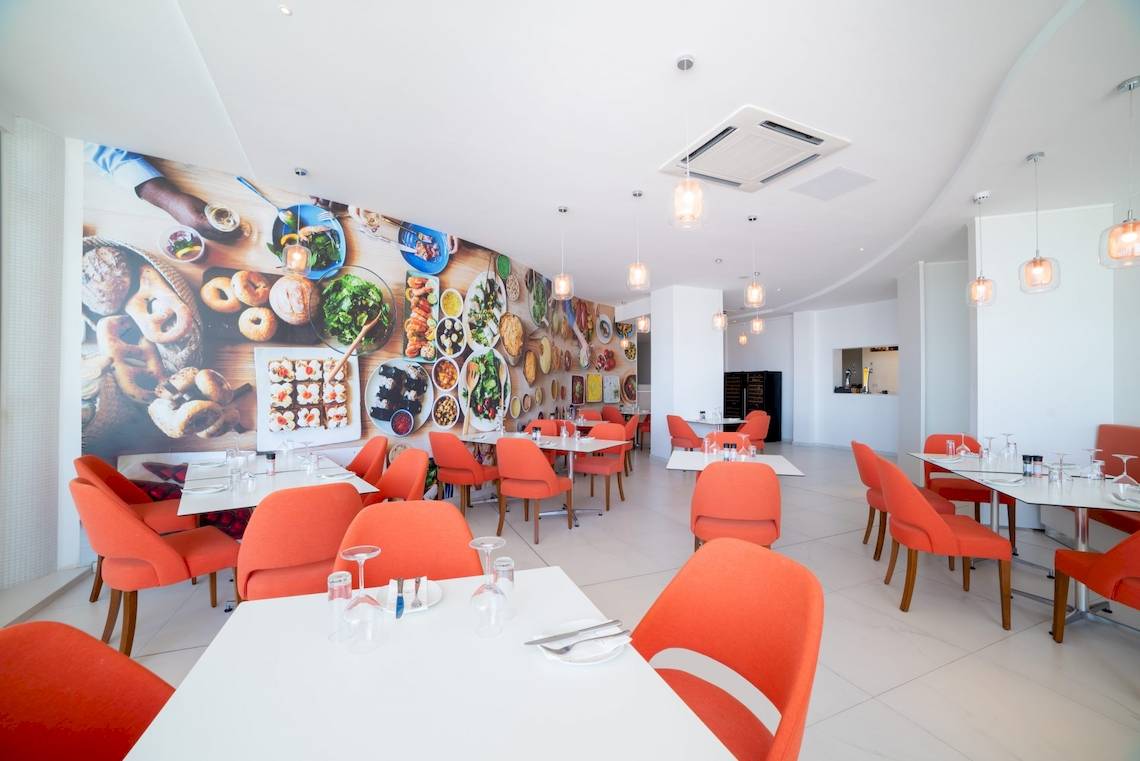 Innenbereich des Ramla Bay Resorts mit orangefarbenen Stühlen und Tischen, einer großen Wandmalerei mit Essensmotiven und modernen Kronleuchtern.