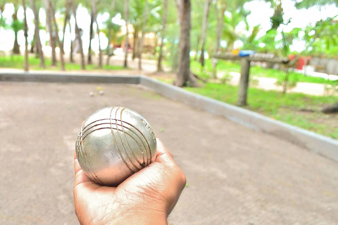 Hand hält einen silbernen Petanque-Ball vor einem Spielfeld mit Bäumen im Hintergrund.