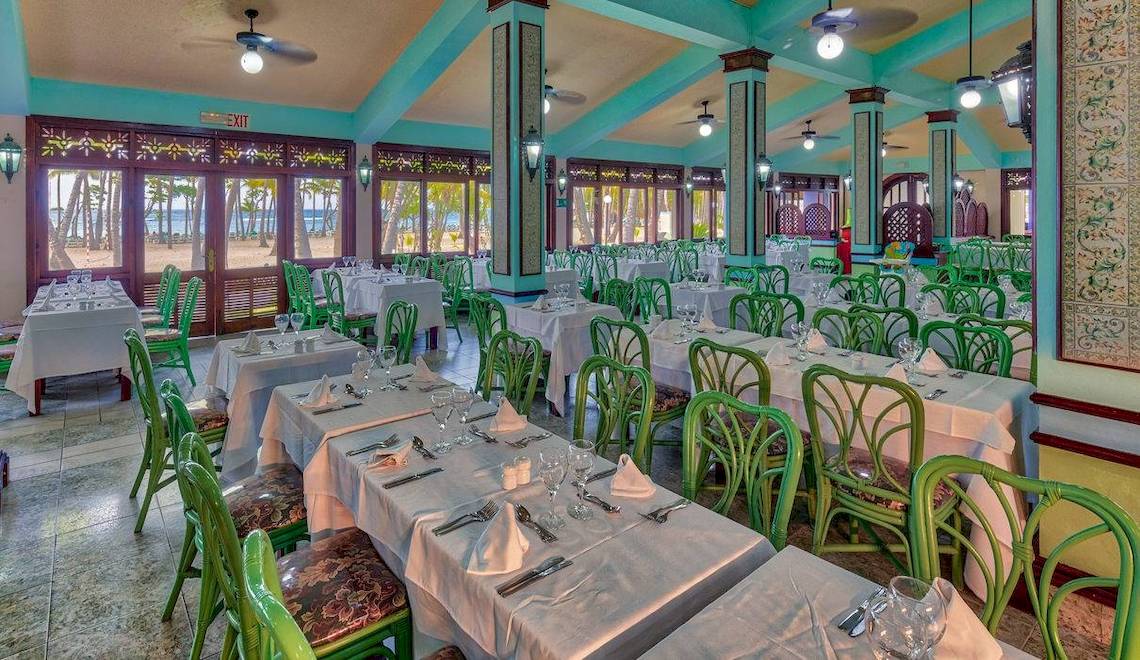 Innenansicht des Restaurants im Playabachata Spa Resort mit türkisblauen Wänden, weißen Tischen und grünen Stühlen, mit Blick auf den Strand.