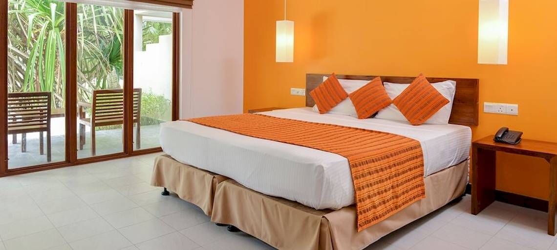 Hotelzimmer im Adaaran Select Hudhuran Fushi mit weißem Bett, orangen Kissen und Tisch neben dem Bett.