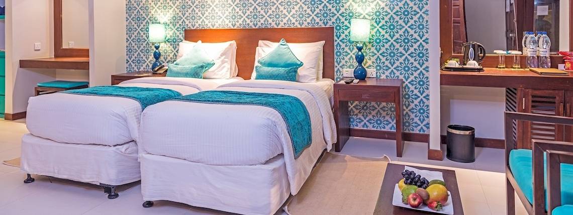 Doppelbettzimmer im Adaaran Select Hudhuran Fushi mit blauen und weißen Musterwänden, zwei Betten, Tischlampen und Obstteller.