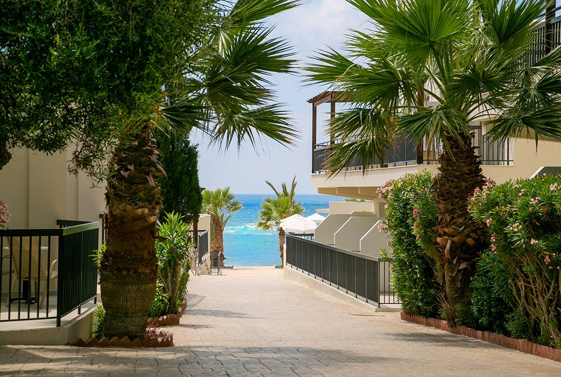 Eingang des Louis Paphos Breeze Hotels mit Blick auf das Meer, umgeben von Palmen und Gebäuden.