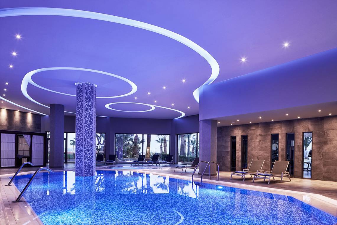 Innenansicht des Schwimmbads im Rodos Princess Beach Hotel mit violetter Beleuchtung und Lounge-Liegestühlen.