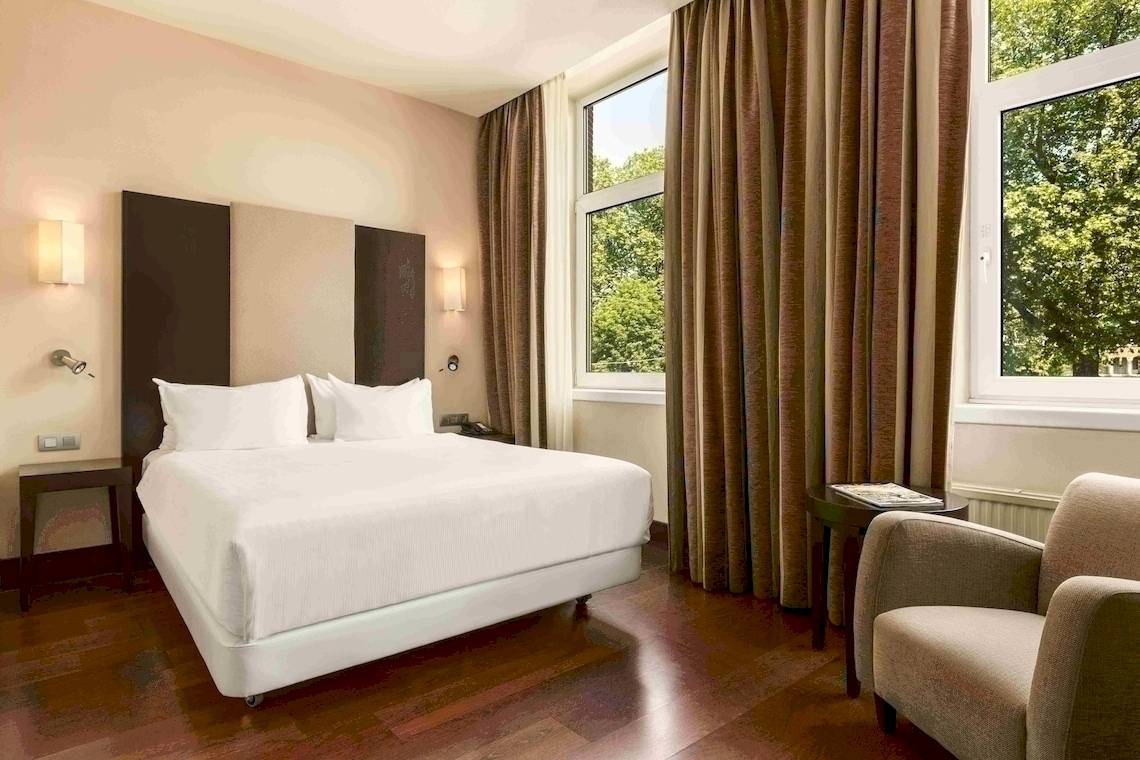Ein elegantes Hotelzimmer im NH Amsterdam Centre mit einem Bett, einem Stuhl und großen Fenstern.