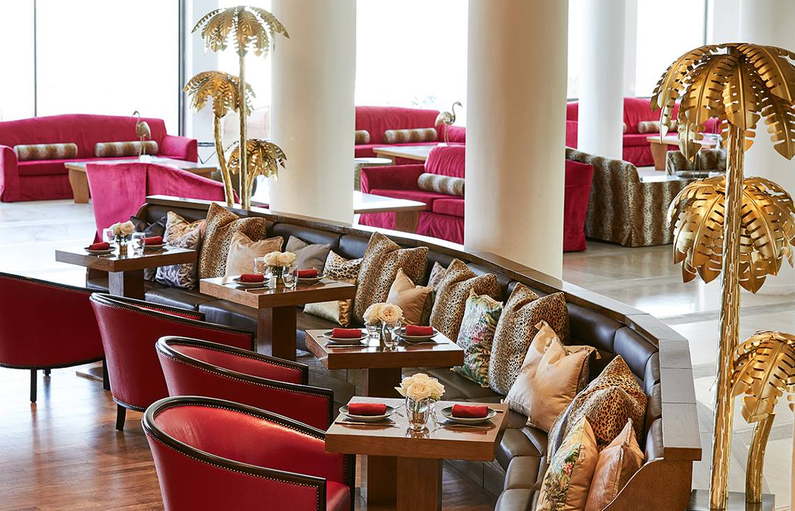 Innenbereich des Grecotel Lux Me Dama Dama Hotels mit roten Möbeln und goldenen Palmen.