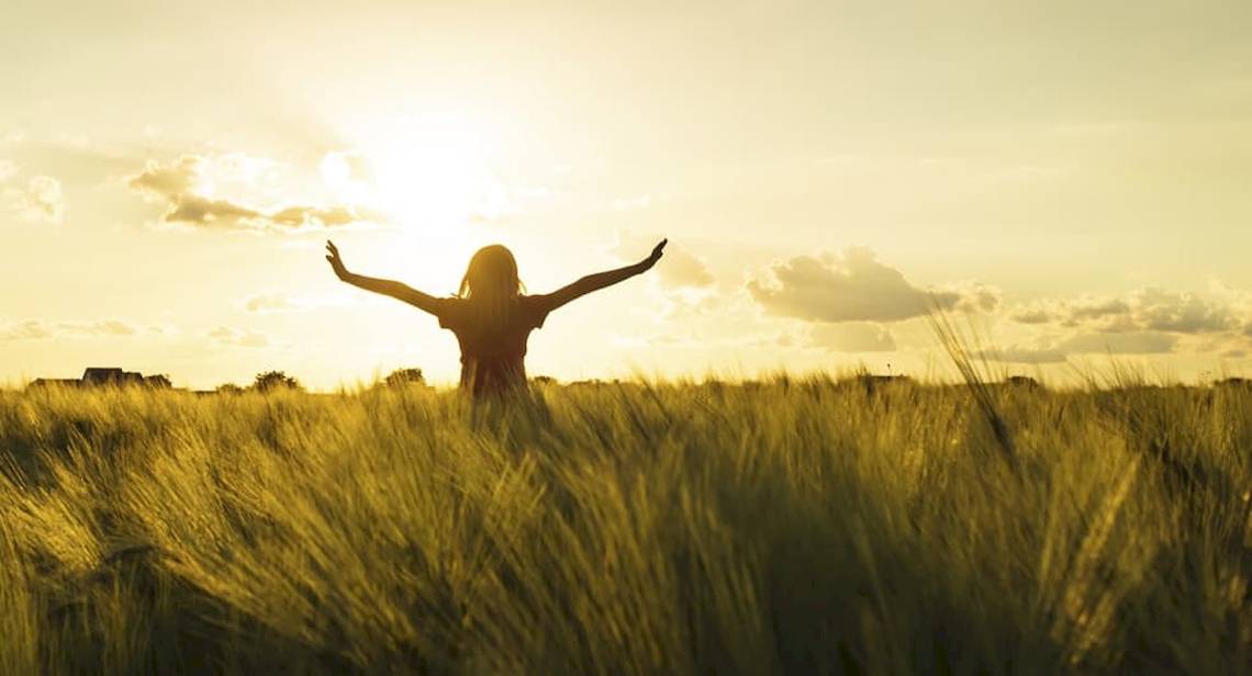 Frau im Feld bei Sonnenuntergang mit ausgebreiteten Armen.
