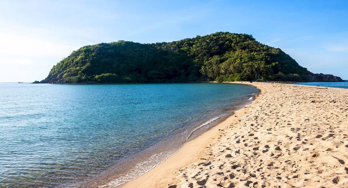 Eine Sandstrand mit klarem Wasser und einem Berg im Hintergrund.
