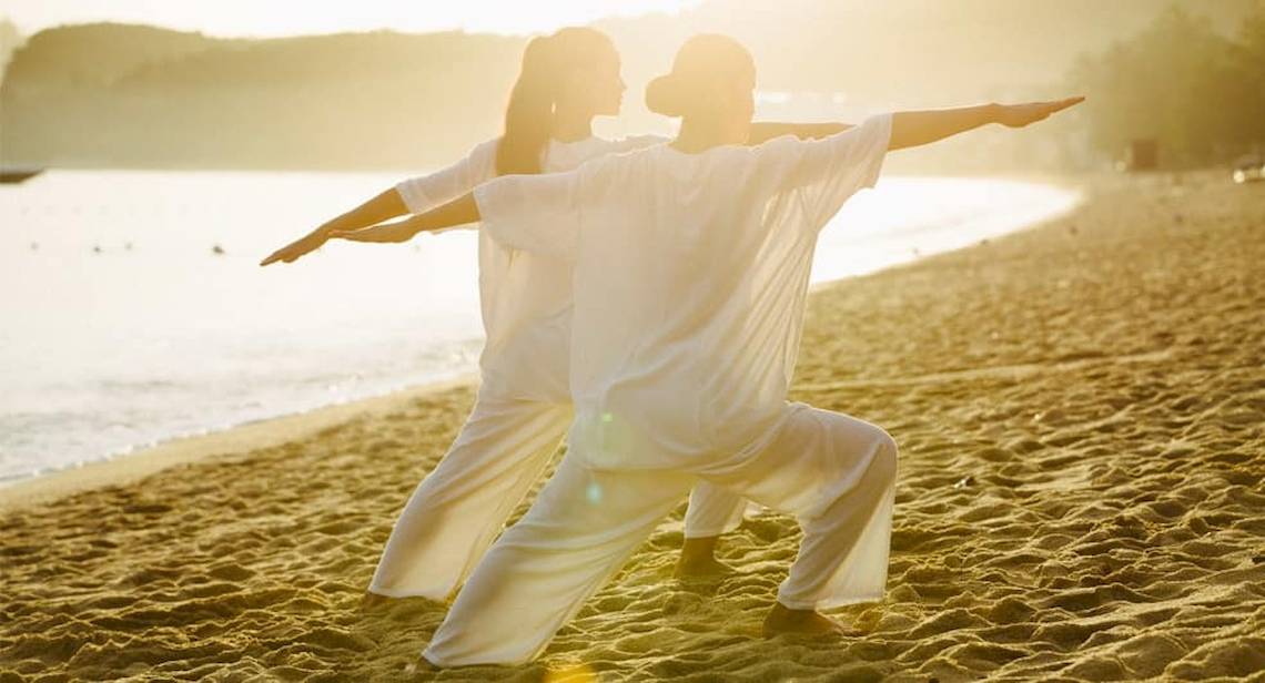 Zwei Personen praktizieren Yoga auf einem Strand bei Sonnenaufgang.