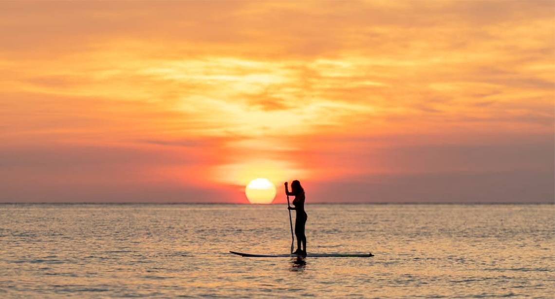 Eine Frau steht auf einem Stand-Up-Paddler bei Sonnenuntergang am Strand