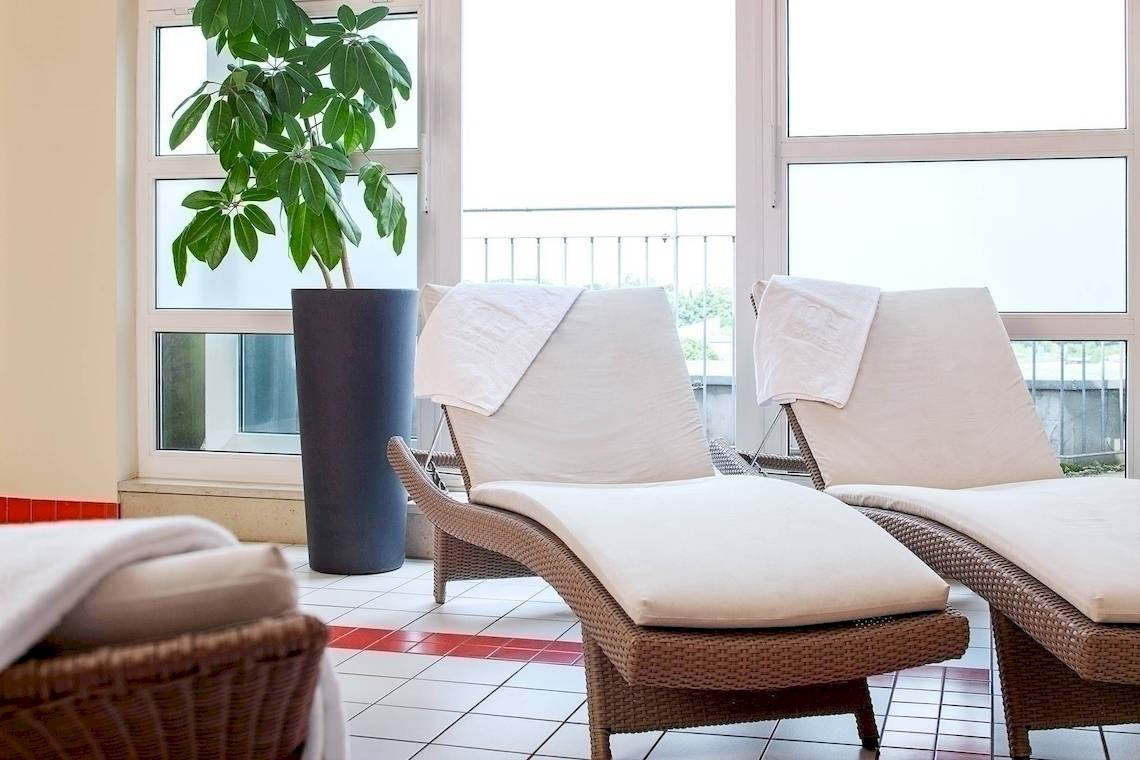 Loungebereich im NH Hamburg Altona Hotel mit zwei weißen Liegestühlen und einem großen Topf mit grüner Pflanze.