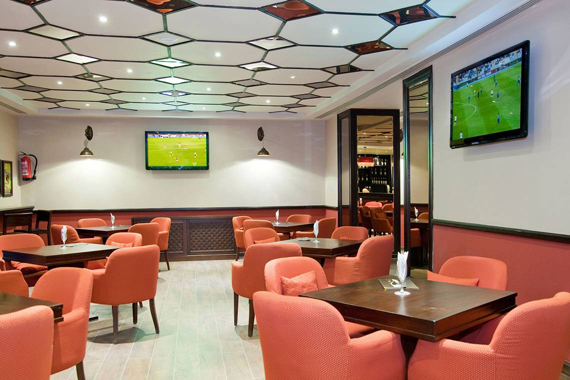 Innenansicht des Restaurants im Grand Waterworld Makadi Hotel mit orangefarbenen Stühlen und Tischen, Fernsehern an der Wand und einer gemusterten Decke.