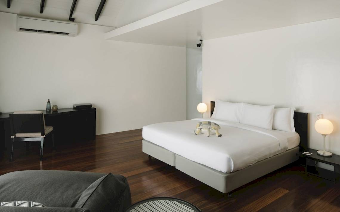 Innenansicht eines Schlafzimmers im Noku Maldives Hotel mit einem Bett, Nachttischen und Schreibtisch.