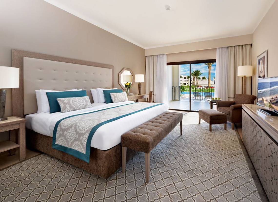 Hotelzimmer im Steigenberger Alcazar mit Kingsize-Bett, gepolsterter Bank, zwei Sesseln und Fernseher.