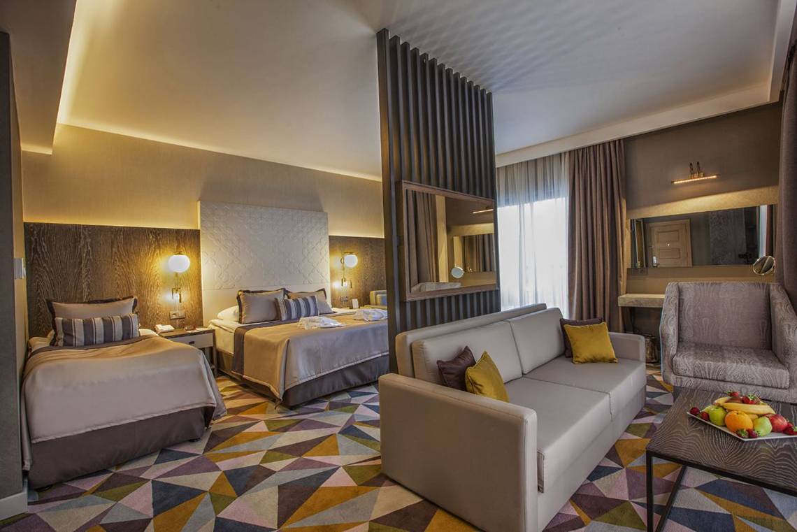 Suite Zimmer im Fame Residence Lara & Spa Hotel mit zwei Betten, Sofa, Tisch und Obstplatte.