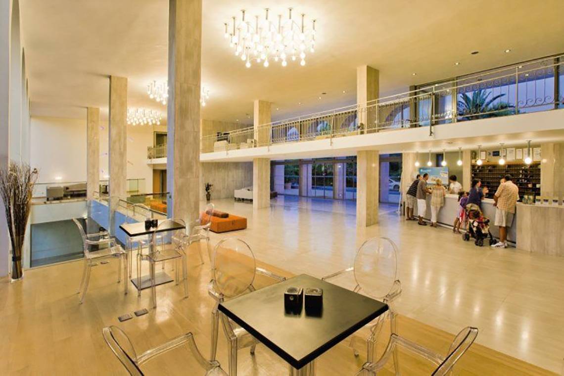 Foyer im Caravia Beach Hotel mit Gästen an der Rezeption und Tischen im Erdgeschoss.