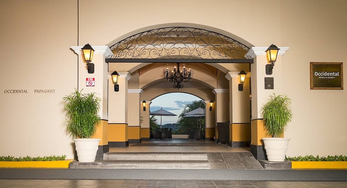 Die Vorderansicht des Occidental Papagayo Hotels mit einer gebogenen Eingangstür, einem Kronleuchter, zwei großen Pflanzen in weißen Töpfen und einem gepflasterten Eingang.