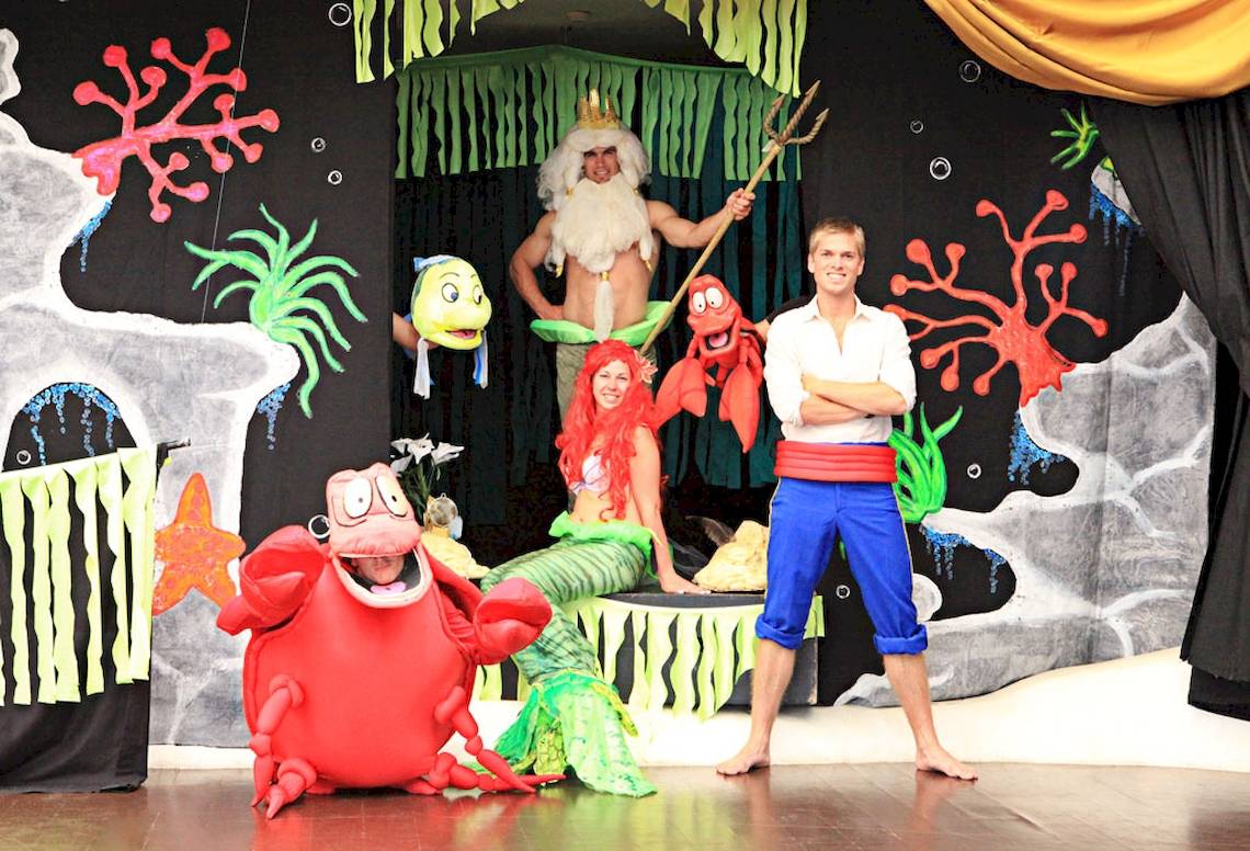 Vier Personen in Kostümen auf einer Bühne, zwei als Arielle und Sebastian, einer als König Triton und einer als Mann in einem weißen Hemd.