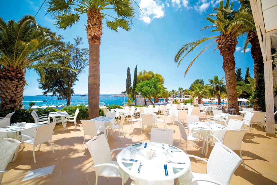 Außenbereich des LABRANDA TMT Bodrum Hotels mit vielen weißen Tischen und Stühlen, umgeben von Palmen und Blick auf das Meer.