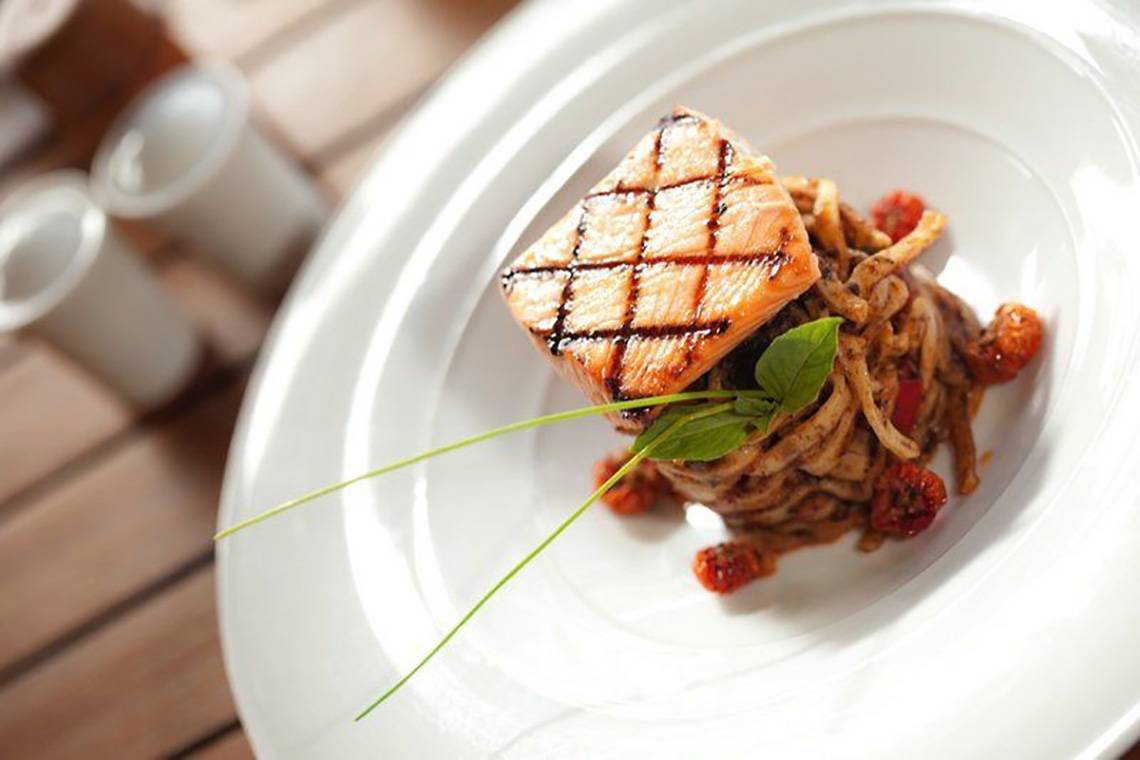 Grillierter Lachs auf Nudeln mit Tomaten und Basilikum auf einem weißen Teller.