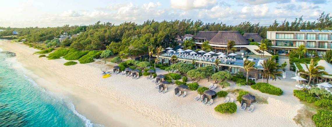 Außenansicht des Recif Attitude Hotels mit Strand, Bäumen, Sonnenschirmen und Lounge-Stühlen.