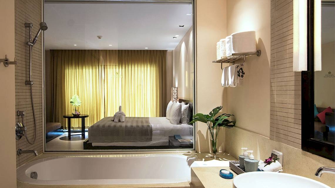 Badezimmer im Apsara Beachfront Resort und Villa mit Badewanne, Spiegel und Bett im Hintergrund.