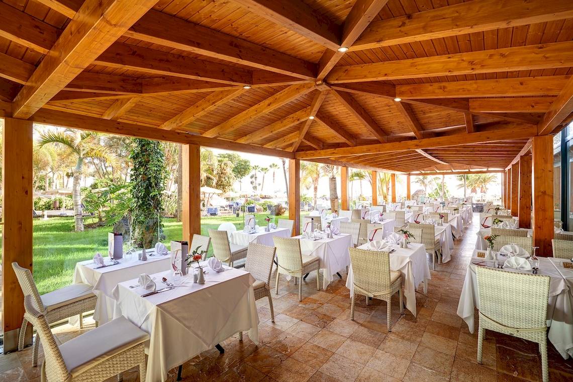 Innenbereich des Restaurants im Hipotels Natura Palace mit Holzbalken und grüner Rasenlandschaft im Hintergrund.