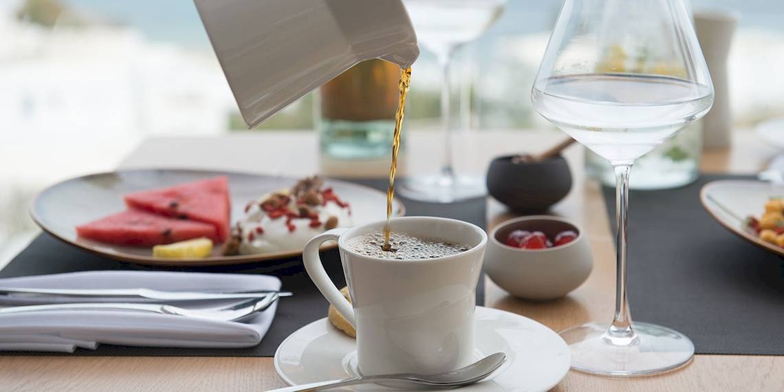 Frühstückstisch im Hotel Myconian Korali Relais & Chateaux mit Kaffee, Wasserglas und Teller mit Wassermelonen, Joghurt und anderen Speisen.