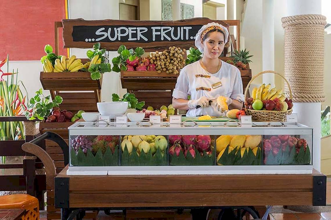 Frau am Cape Panwa Hotel, die eine Ananas schneidet, mit einem Obststand im Hintergrund.