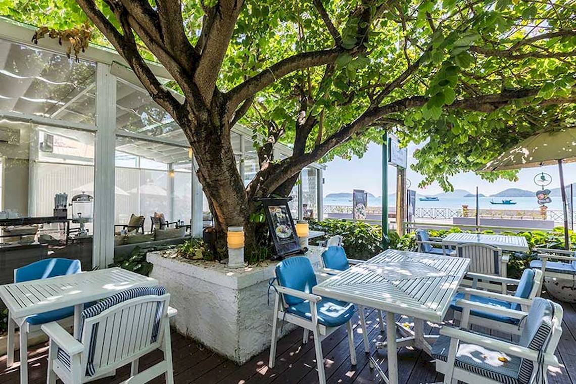 Außenterrasse des Cape Panwa Hotels mit Tischen, Stühlen und einem großen Baum