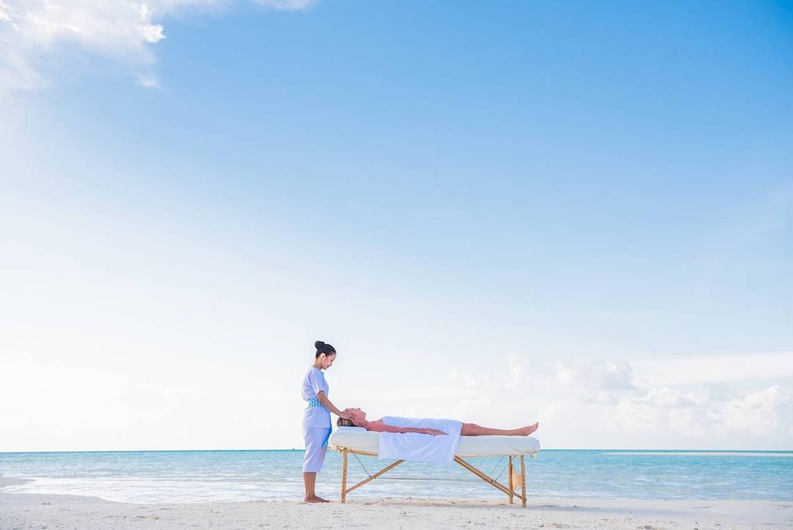 Eine Massage an einem Strand, durchgeführt von einem Therapeuten, mit einem Klienten, der auf einer Massageliege liegt, im Kandima Maldives Hotel.