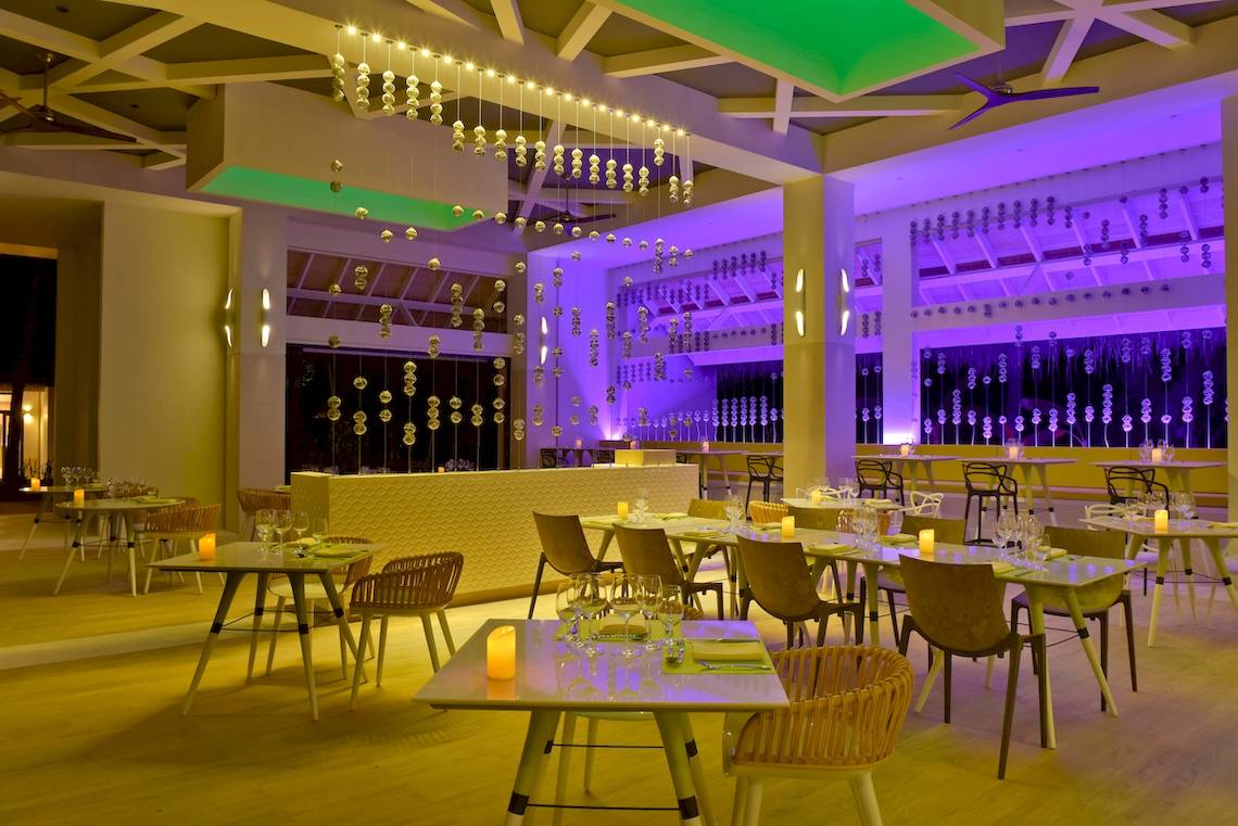Innenansicht des Restaurants im Kandima Maldives Hotel mit runden Tischen, Stühlen und hängenden Lichtern.