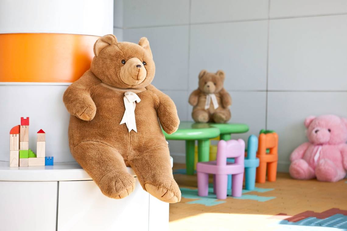 Ein großer brauner Teddybär sitzt auf einem weißen Schrank im Kinderzimmer neben bunten Spielzeugen.
