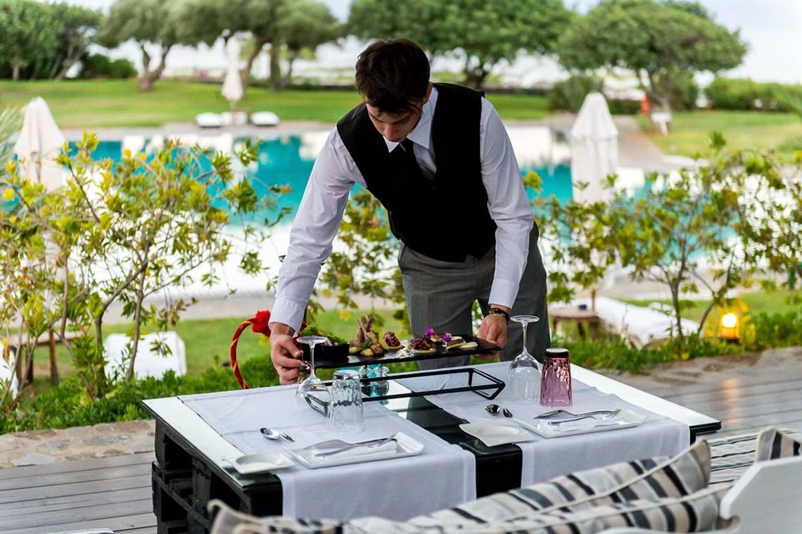 Ein Kellner serviert ein Tablett mit Speisen auf einer Terrasse im Elounda Beach Hotel & Villas.