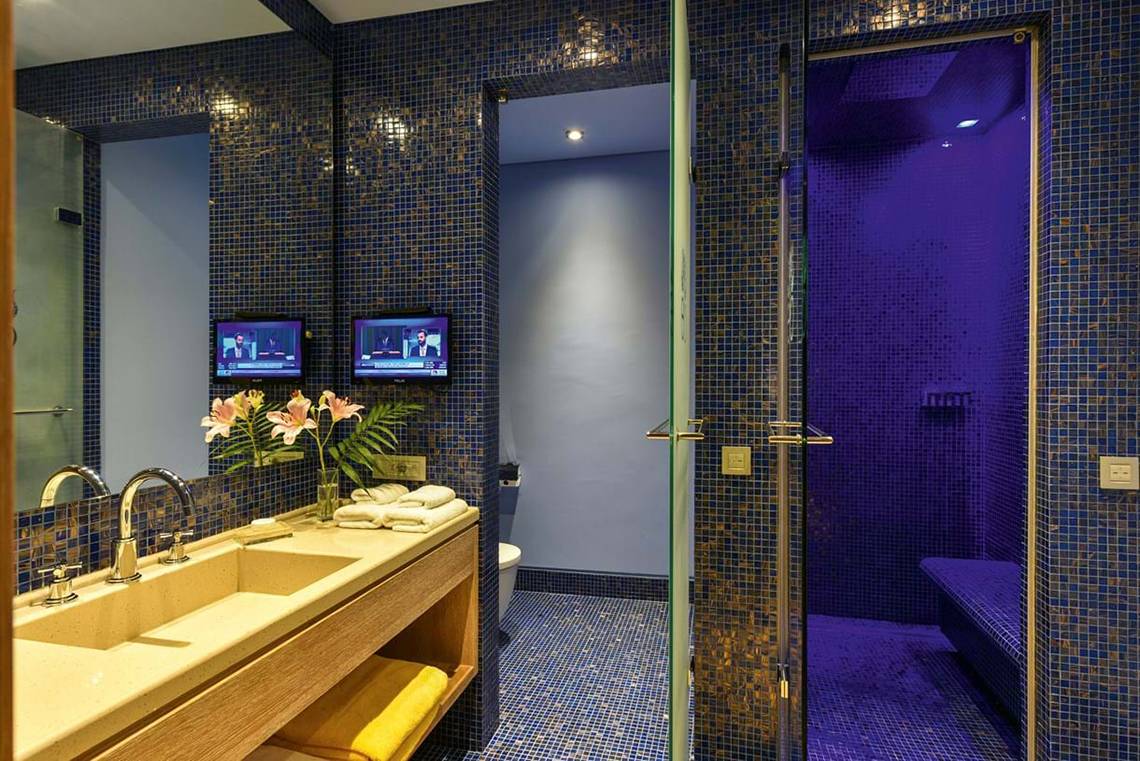 Badezimmer im Elounda Beach Hotel & Villas mit blauen Kacheln, doppeltem Waschbecken und Fernsehern an der Wand.