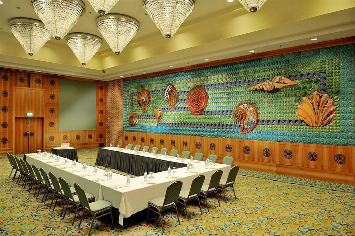 Konferenzraum im Gloria Verde Resort mit großen Tisch, farbigen Wanddekorationen und Kronleuchtern.