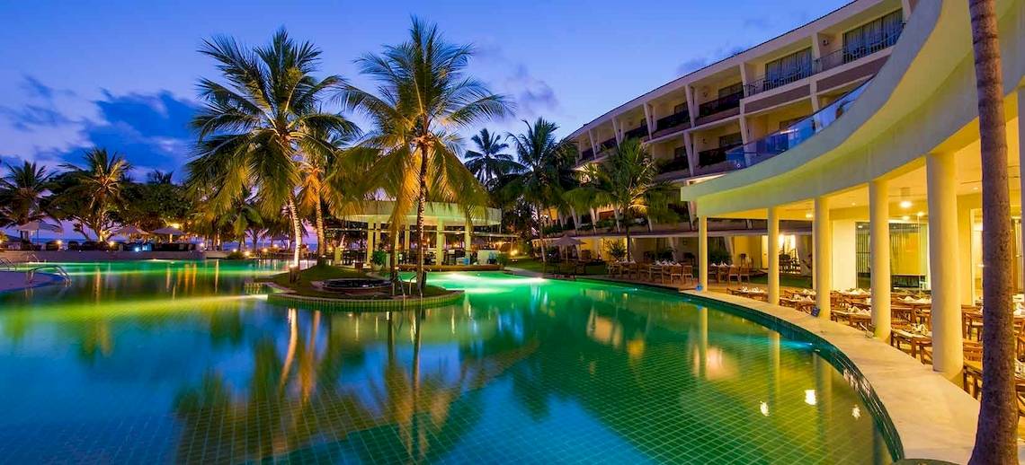 Ein großer Swimmingpool mit leuchtend grünem Wasser, umgeben von Palmen, vor dem Occidental Eden Beruwala Hotel bei Nacht.