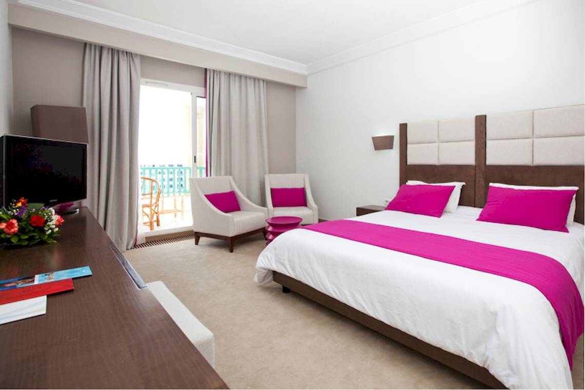 Hotelzimmer im El Mouradi Hammamet mit großem Bett, zwei Sesseln, Fernseher und Balkon.