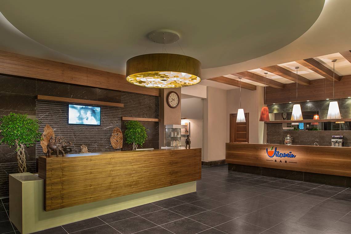 Rezeption im Aska Lara Resort & Spa mit Holzrezeptionstisch, Pflanzen und Bar.
